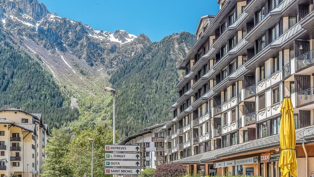 A hotel with multiple balconies, a mountain backdrop, and directional signs for nearby locations. - Appartement Courmayeur - Welkeys (Chamonix-Mont-Blanc)