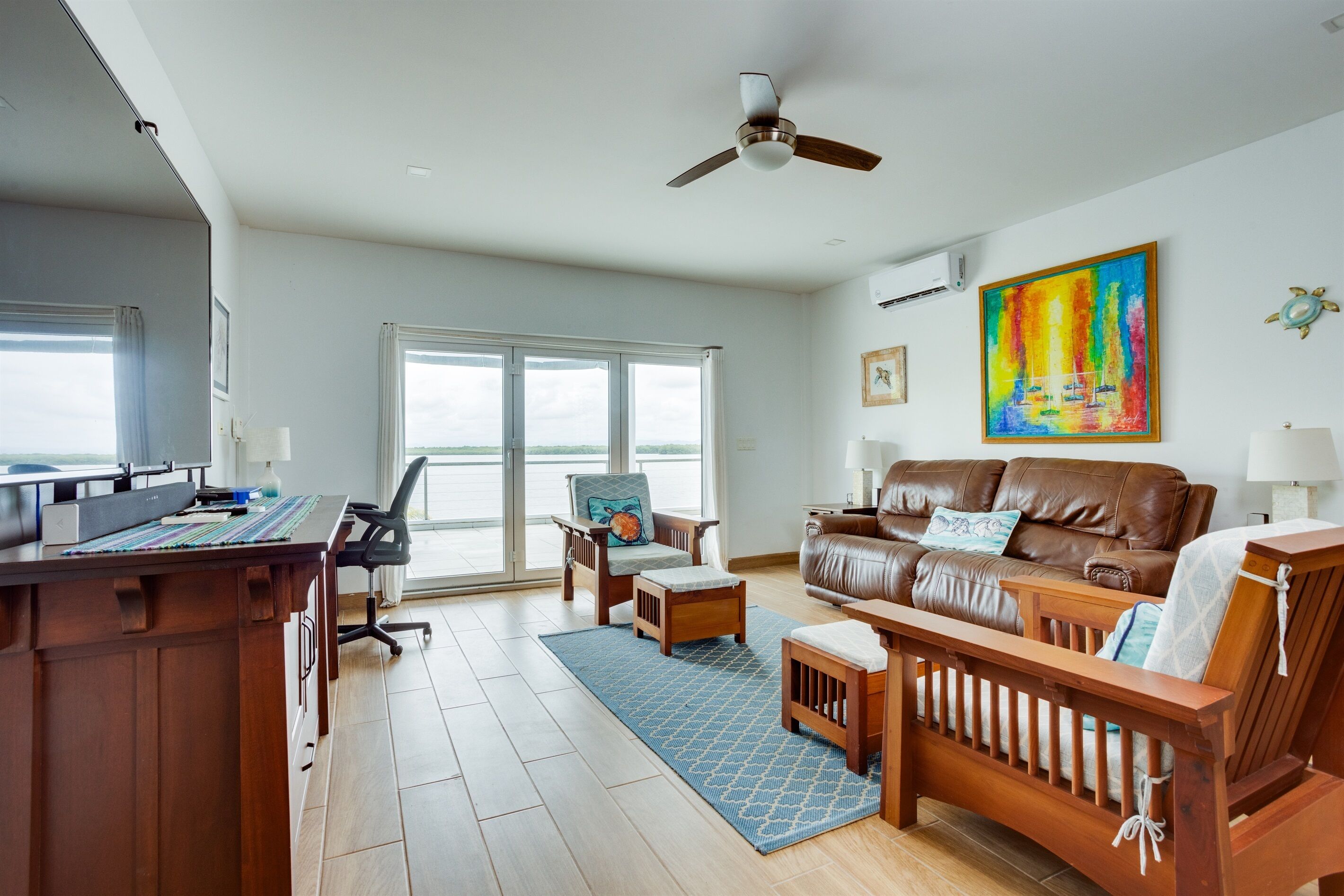 A spacious living room with a brown leather sofa, a wooden coffee table, and a desk with a computer. There is a large window providing a view of the outdoors.