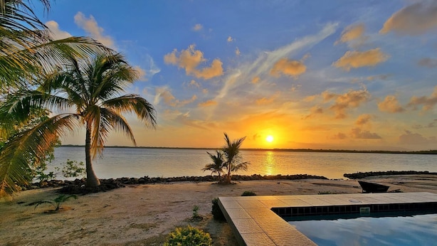 Un área de piscina con vista al atardecer, palmeras y un mar tranquilo.