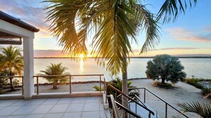 A terrace with a view of the sea, palm trees, and a sunset.