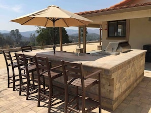 An outdoor bar area with a stone counter, wooden chairs, and a large umbrella.