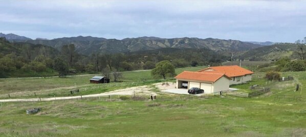 A rural house with a red roof, surrounded by a green landscape and mountains in the background.