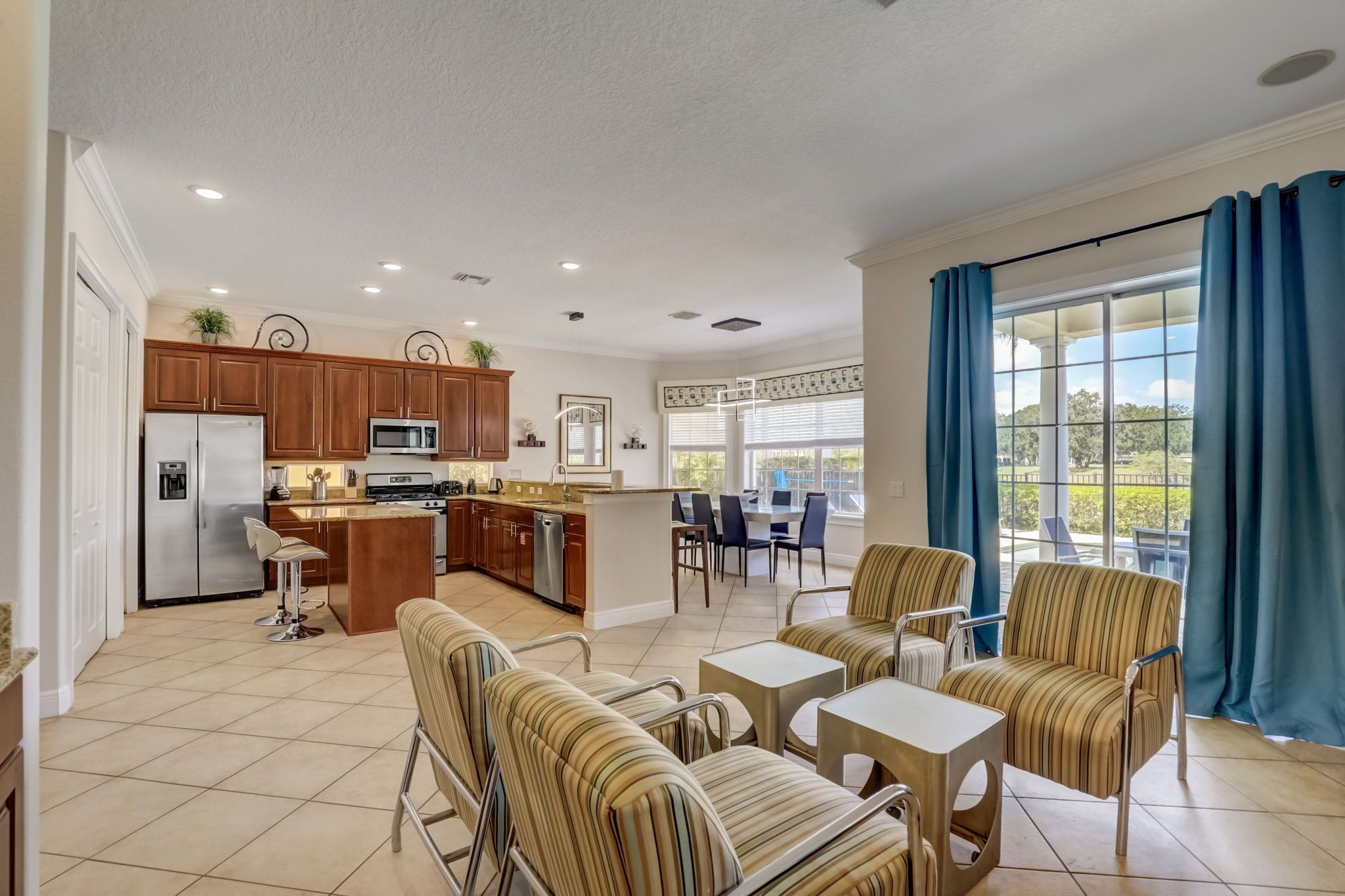 A spacious kitchen with wooden cabinets and a central island, a dining area with striped chairs, and a large window with blue curtains.