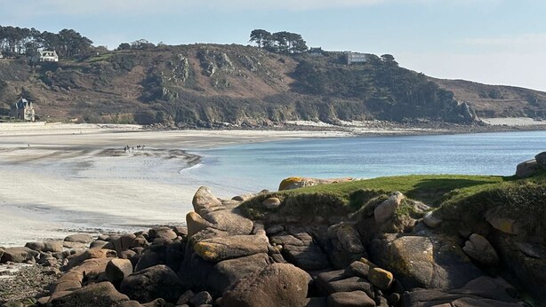 Eine Küstenlandschaft mit felsiger Küste, einem Sandstrand und einem Hügel mit Gebäuden.