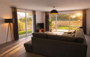 Ein modernes Wohnzimmer mit einer Couch, einem Fernseher und einem großen Schiebeglasfenster, das auf eine Terrasse mit Tisch und Stühlen führt.