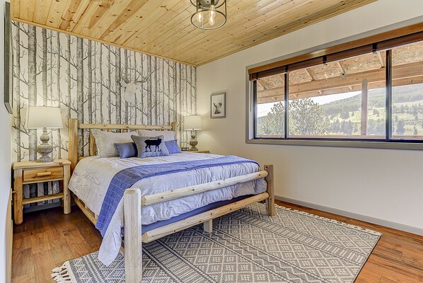 A bedroom with a bed, wooden headboard, nightstands, a window with a view, and a patterned rug. - Monarch Landing, A Cozy Cabin w/360 Mountain Views (Cripple Creek)