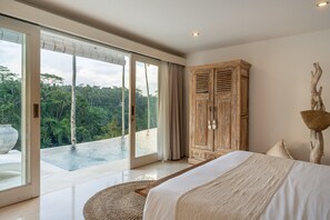 A bedroom with a large bed, a wooden wardrobe, and a view of a natural landscape through a sliding glass door. - Sacred Jungle • Luxury 4BR Ubud • 2 Infinity Pools (Ubud)