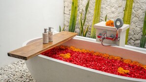 Une baignoire remplie de pétales de fleurs aux couleurs vives, une étagère en bois avec des articles de toilette et un mur de pierre orné de plantes vertes.