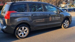 Een grijze auto met de branding van 'La Baulé Hôtel de Charme' geparkeerd voor een historisch gebouw met een klokkentoren.