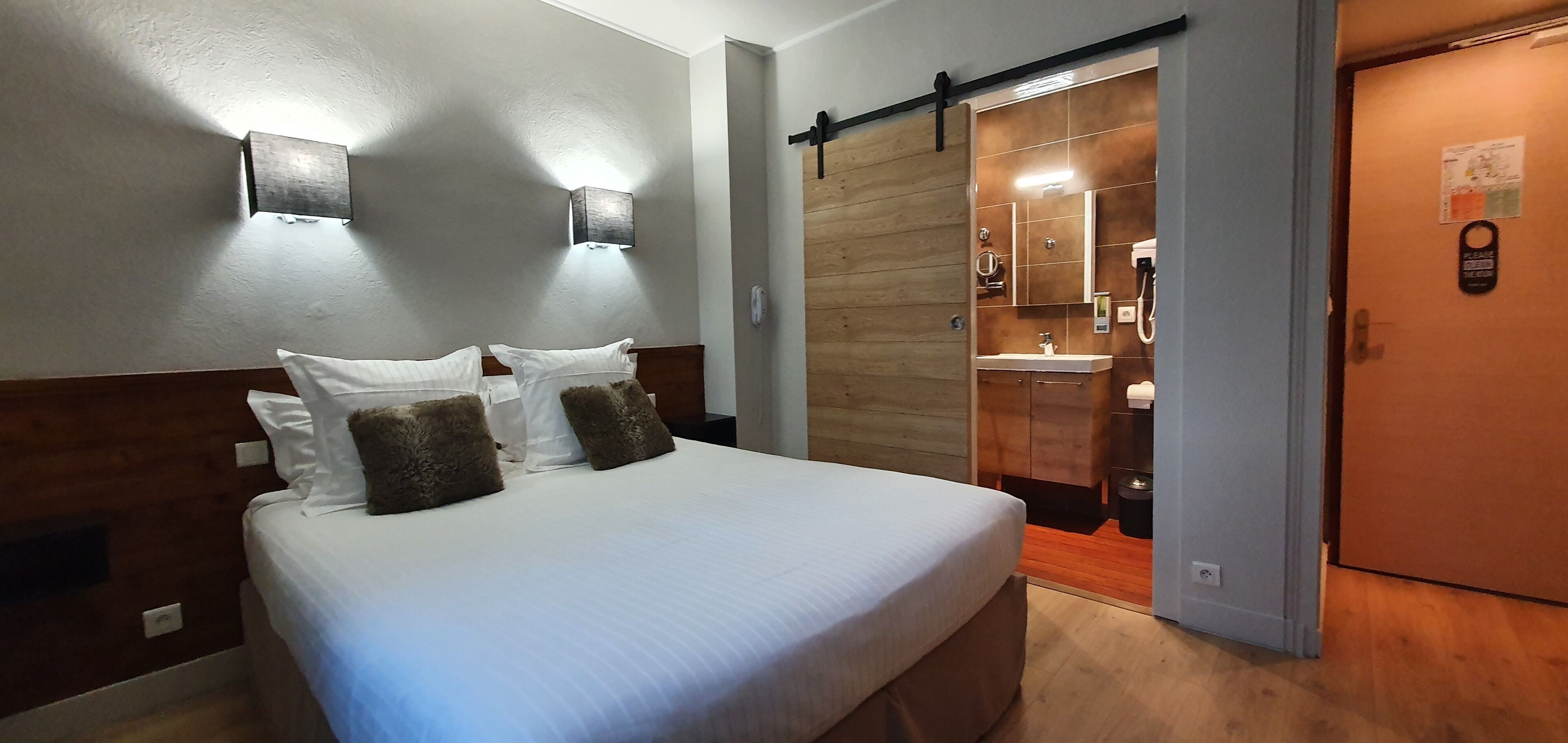 Une chambre à coucher comprenant un lit, deux lampes fixées au mur, une tête de lit en bois et une porte coulissante donnant sur une salle de bain.