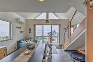 A modern living room with a large window, a sofa, a dining table, and a staircase leading to an upper level. - Yachats Gem w/ Private Beach Access, Views (Yachats)
