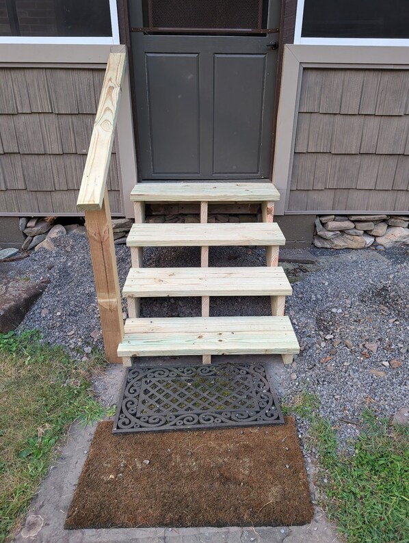 A wooden outdoor staircase leading to a door, with a doormat and gravel path in front. - Vacation Property off the grid. No cell service, internet, satellite/ cable tv (Renovo)