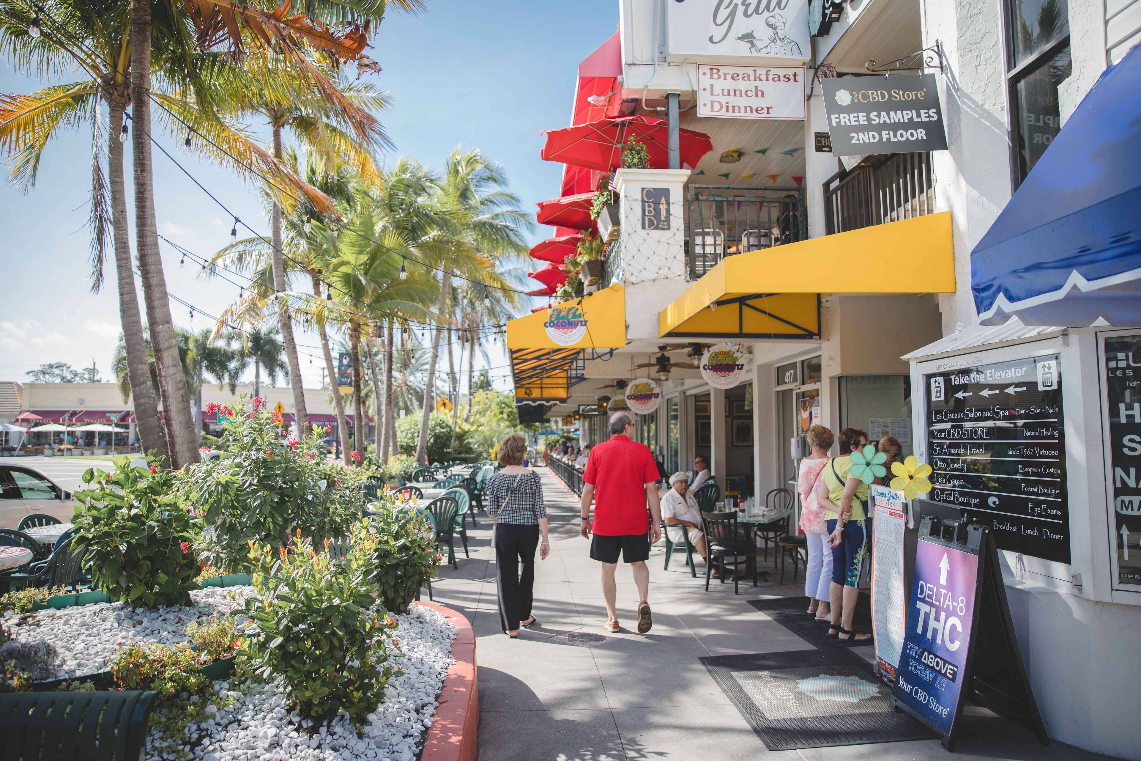 Une rue bordée de terrasses, de palmiers et d’un magasin de produits à base de CBD qui offre des échantillons gratuits au deuxième étage.