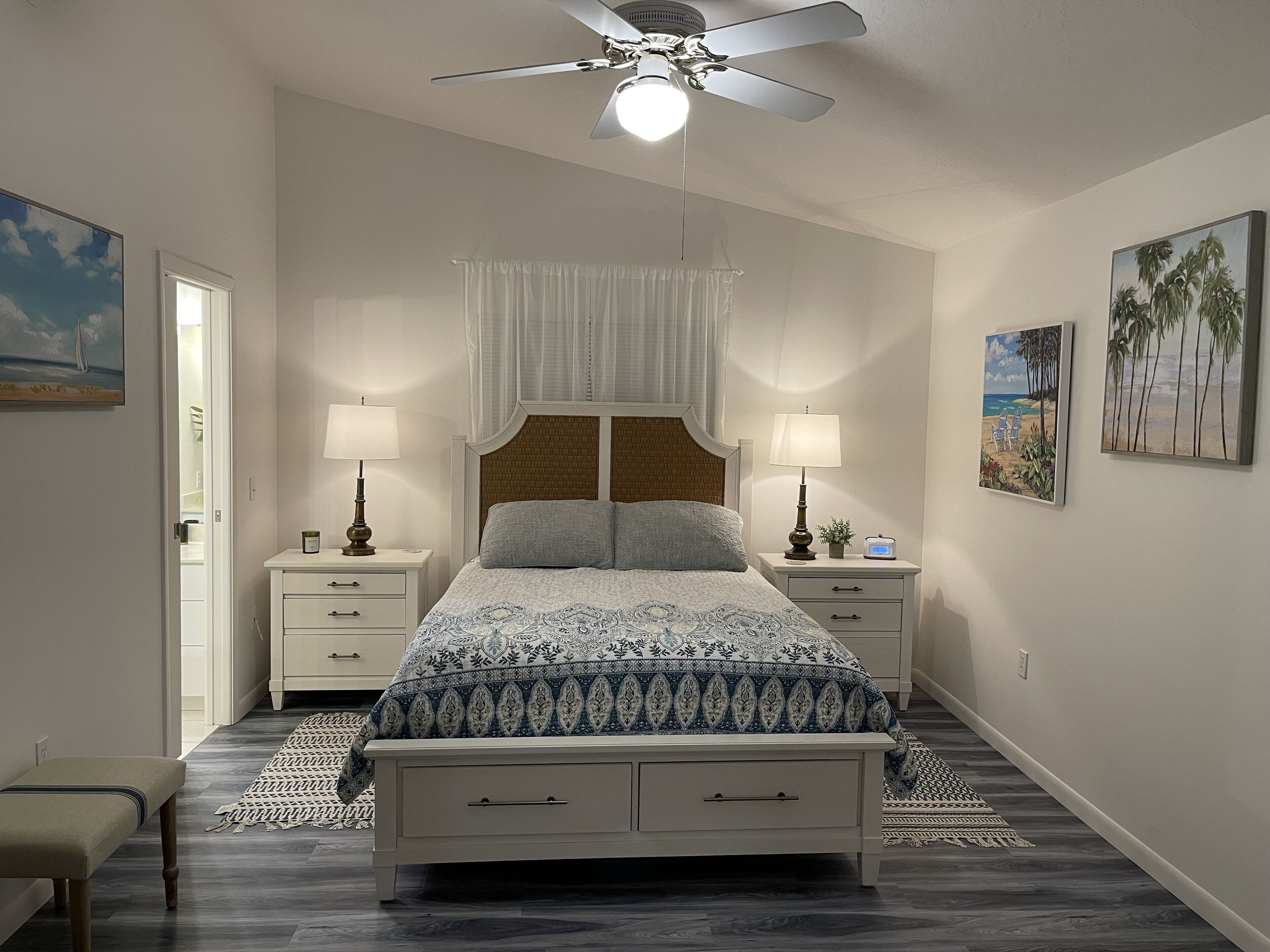 Une chambre à coucher comprenant un lit, deux tables de chevet, un ventilateur de plafond et des œuvres d’art accrochées aux murs.