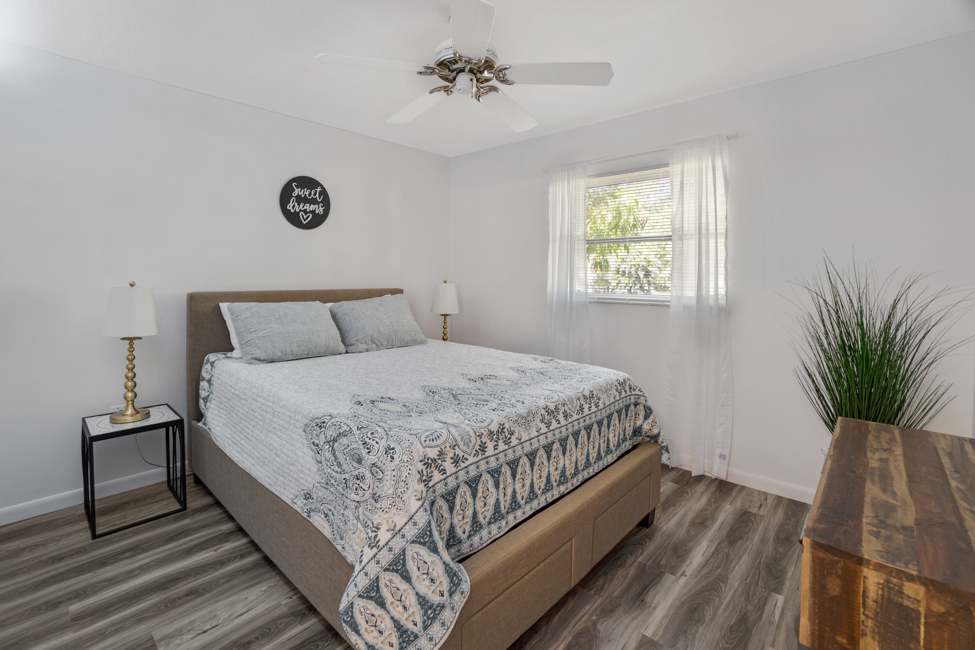 A bedroom with a bed, bedside lamps, a ceiling fan, a window with curtains, and a plant.