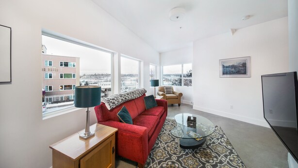 A modern living room with a red sofa, a glass coffee table, and a view of the city through large windows. - Stylish Downtown Loft 201-Alaska Railroad Suite (Anchorage)