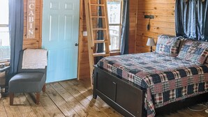 Uma cabana acolhedora com um quarto no mezanino, uma escada para o espaço de dormir superior, uma cama com edredom xadrez e uma poltrona com manta.