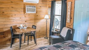 Interior de uma cabana com piso de madeira, uma cama com colcha xadrez, uma mesa de jantar com cadeiras, um abajur e uma janela com cortinas.