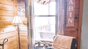 Interior de cabana com piso de madeira, uma cadeira com toalha, uma luminária e janela com vista para um edifício.