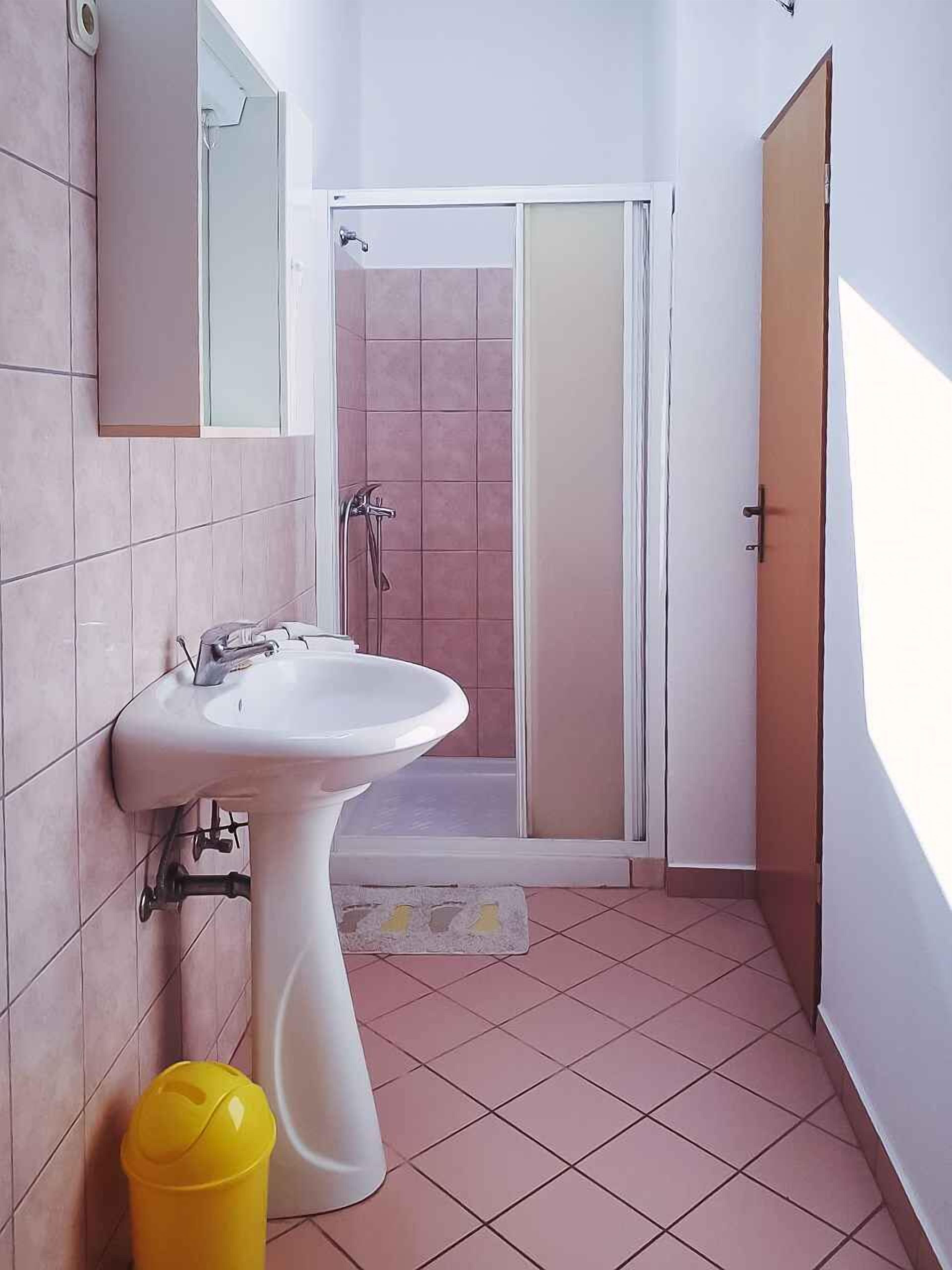 A bathroom with a pedestal sink, a shower with a glass door, and a yellow trash bin.