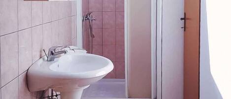 A bathroom with a pedestal sink, a shower with a glass door, and a yellow trash bin.