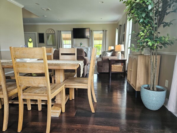 A dining area with wooden chairs and a table, a sofa, a television, and a potted plant.
