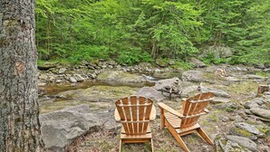 Two wooden Adirondack chairs near a rocky stream in a forested area. - Beautiful Jay Peak Home Near Creek & Waterfalls! (Jay)