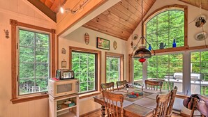 A wooden dining area with a table and chairs, large windows with a view of greenery, and a microwave on a shelf. - Beautiful Jay Peak Home Near Creek & Waterfalls! (Jay)