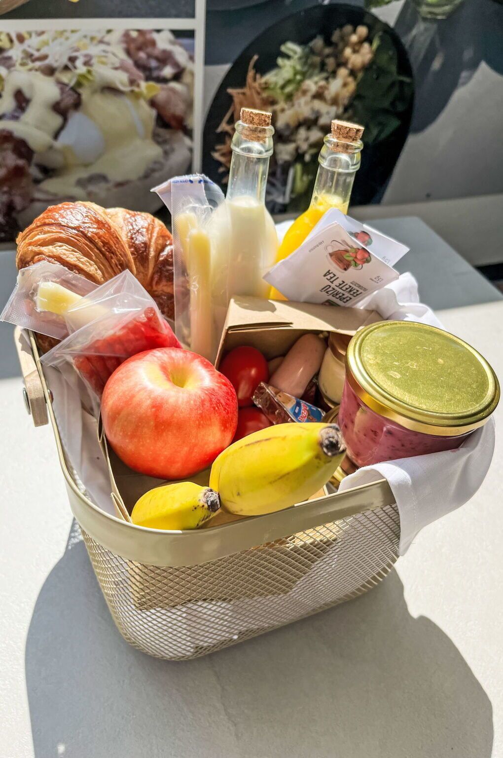 Ein Korb mit einem Croissant, Äpfeln, Bananen, einer Marmeladenglas und Milch- und Saftflaschen.