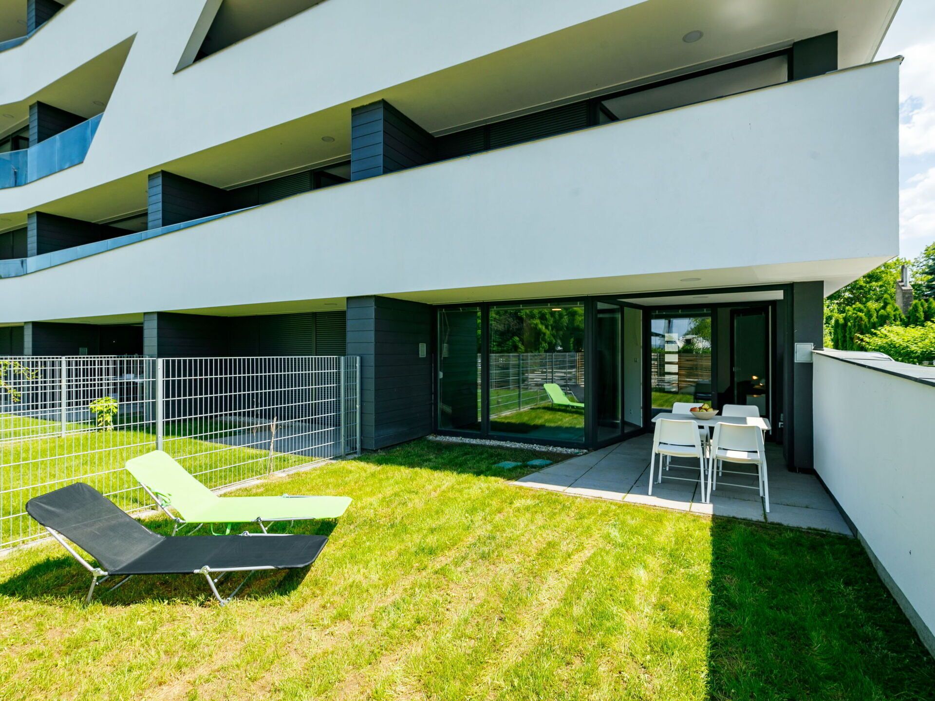 Ein modernes, zweistöckiges Gebäude mit flachem Dach, einer überdachten Außenbereich-Sitzgelegenheit mit weißen Tischen und Stühlen sowie einem eingezäunten Garten mit Rasen und zwei Liegestühlen.