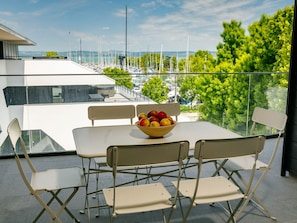 Un balcon avec une table et des chaises, un saladier rempli de fruits et une vue sur les bateaux et la verdure.