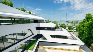 Un edificio moderno con fachada de vidrio, varios niveles y un área ajardinada con árboles y arbustos.