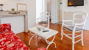 A living room with a red floral sofa, white chairs, a white table with a remote control, a fireplace, and a TV mounted on the wall.