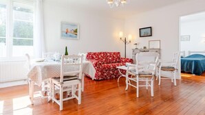 A room with a wooden floor, white furniture, a red floral sofa, a dining table with chairs, and a chandelier.