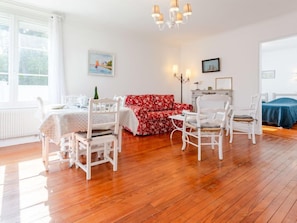 Um quarto com piso de madeira, mobiliário branco, um sofá floral vermelho, uma mesa de jantar com cadeiras e um candelabro.