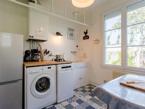 Cocina compacta con lavadora, cafetera y estufa. Hay una ventana con vistas a árboles y una cama con colcha y tabla de madera.