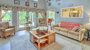 A spacious living room with a large window, a wooden coffee table, a beige sofa, and a kitchen area visible through an open door. - 10-Acre Mountain Home Getaway w/ Fire Pit (Mountain Home)