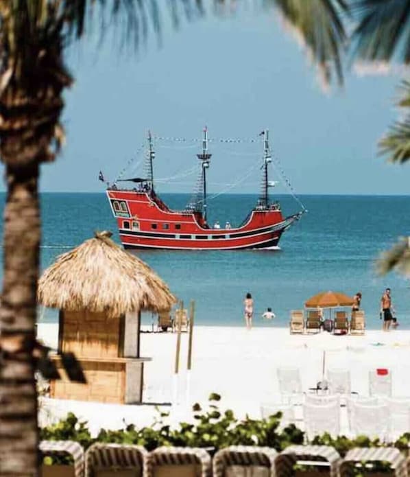 Una playa con un bote rojo en el agua, una cabaña con techo de paja y sillas de playa.