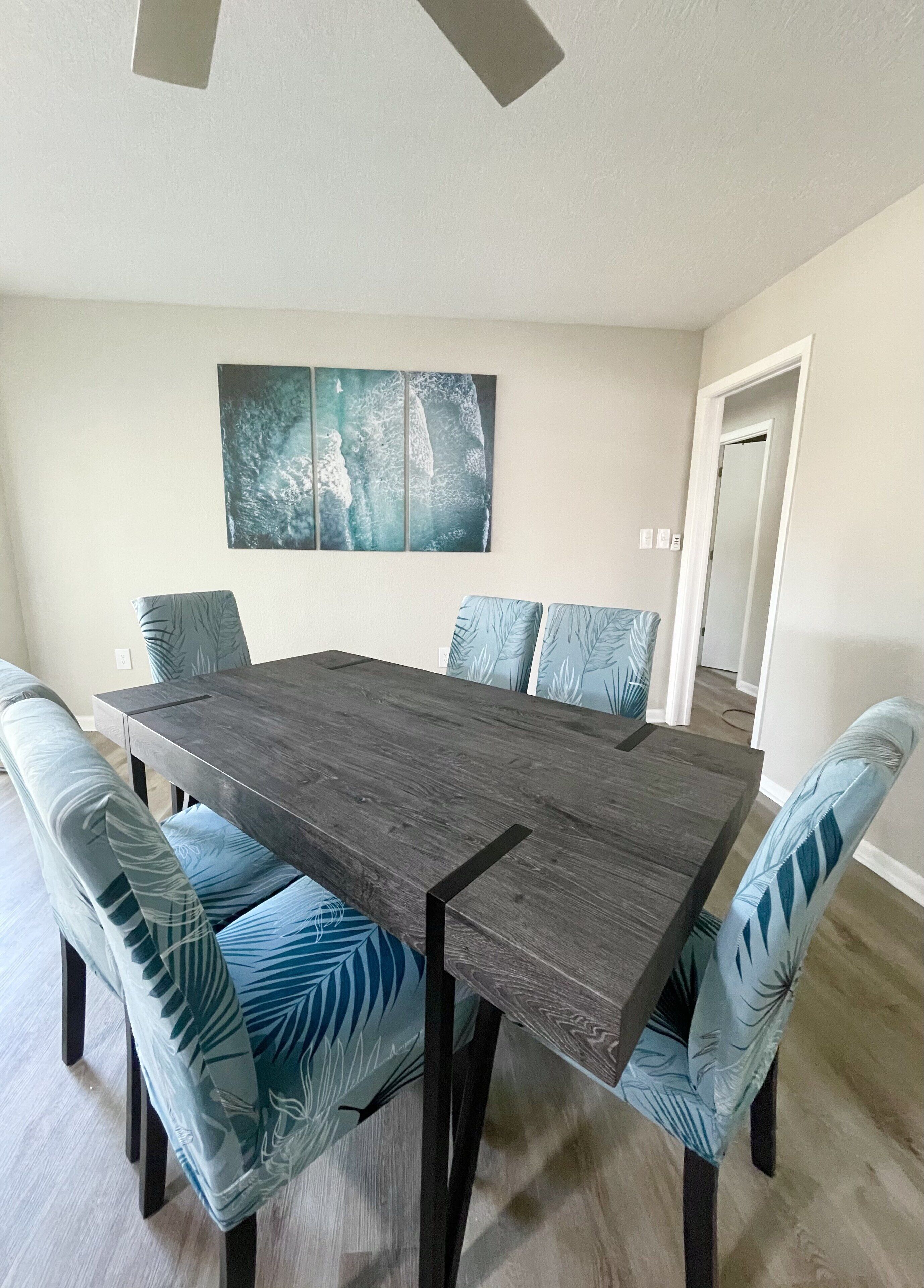 A dining room with a dark wooden table and matching chairs, a large abstract painting on the wall, and a door leading to another room.