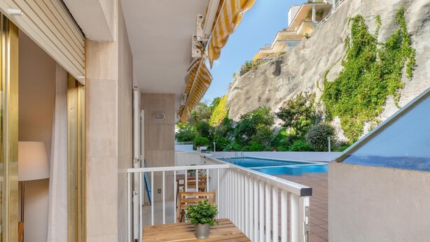 A balcony with a wooden table and chairs, a potted plant, and a view of a swimming pool and greenery.