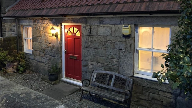 A stone cottage with a red door, a bench outside, and two lit windows.
