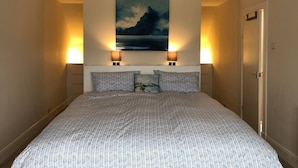 A bedroom with a large bed, two wall-mounted lamps, a painting of a landscape, and a door to the right.