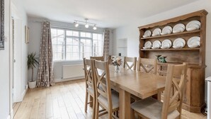 A dining area with a wooden table and chairs, a window with curtains, and a shelf with plates.