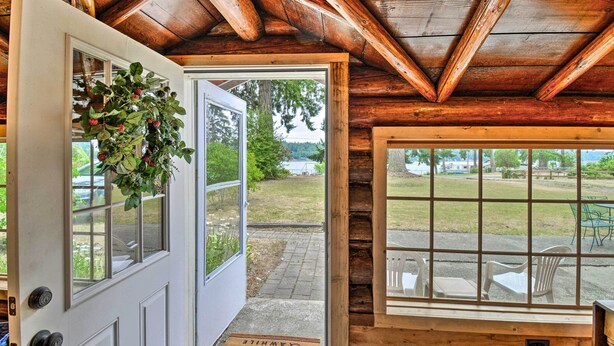 Una baita di legno con una porta che dà su un portico, una finestra con vista su un lago e un tappetino d'ingresso con un messaggio di benvenuto.