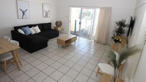 A living room with a black sofa, a wooden coffee table, and a dining area with a table and chairs. There is a ceiling fan, a sliding glass door leading to a balcony, and a TV mounted on the wall.