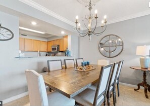 Un comedor con mesa y sillas de madera, una araña y una cocina al fondo.
