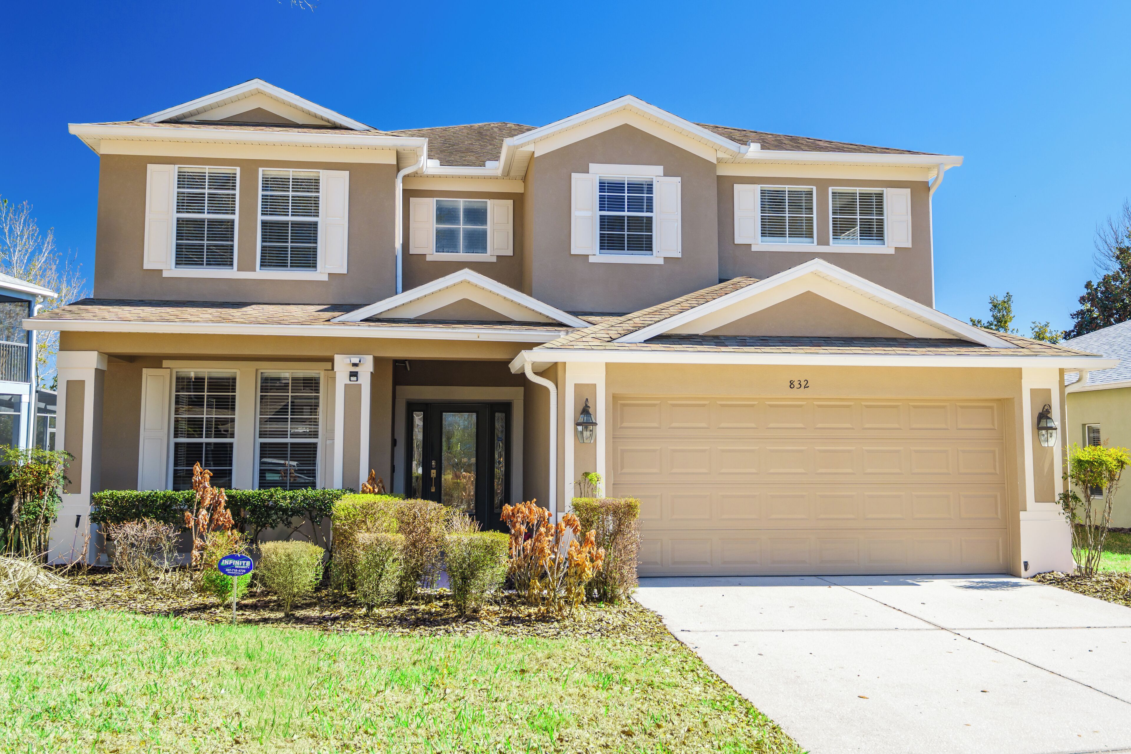 A two-story house with a beige exterior, a garage, and a well-maintained front yard with shrubs and a lawn.