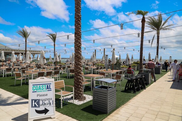 An outdoor seating area with tables and chairs, palm trees, and a sign indicating a coastal location.