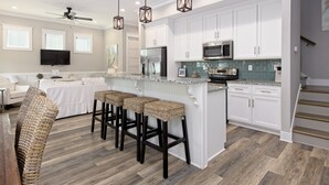 A modern kitchen with white cabinets, a stainless steel refrigerator, a microwave, and a stove. There are three wicker bar stools and a large island with a granite countertop. - Coastal chic beach home in Cottages at Lagoon Pass with EV charger and pool. (Gulf Shores)