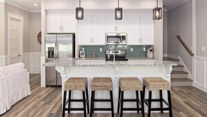 A modern kitchen with white cabinets, stainless steel appliances, and a granite countertop. There are three wicker bar stools and a stainless steel refrigerator. - Coastal chic beach home in Cottages at Lagoon Pass with EV charger and pool. (Gulf Shores)